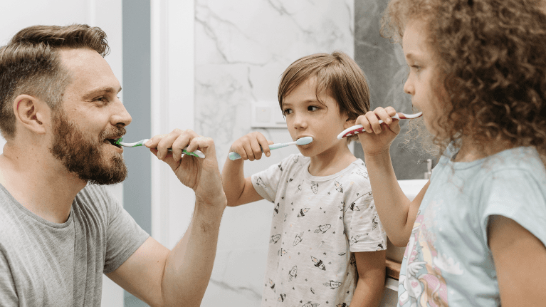 Father and two children brushing their teeth together at home, promoting family dental health and da